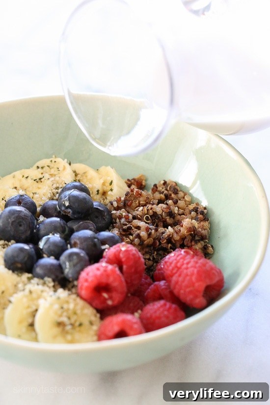 Creamy Berry Quinoa Breakfast Bowl Recipe 2 A bowl with quinoa, sliced bananas, fresh raspberries, fresh blueberries topped with hemp seeds and a creamy honey drizzle being poured on top