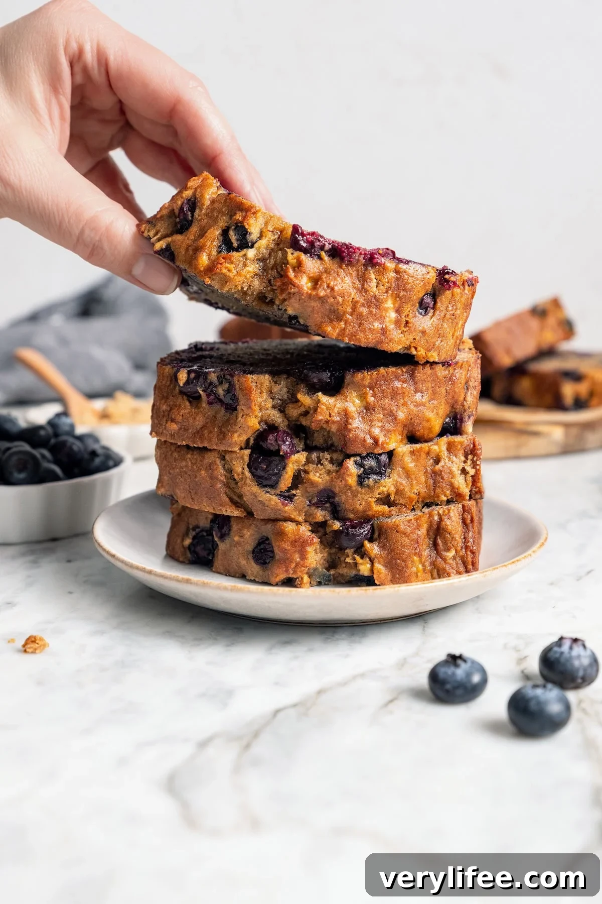 Moist Blueberry Banana Bread Recipe 7 Lifting slice of blueberry banana bread from plate with stack of slices.