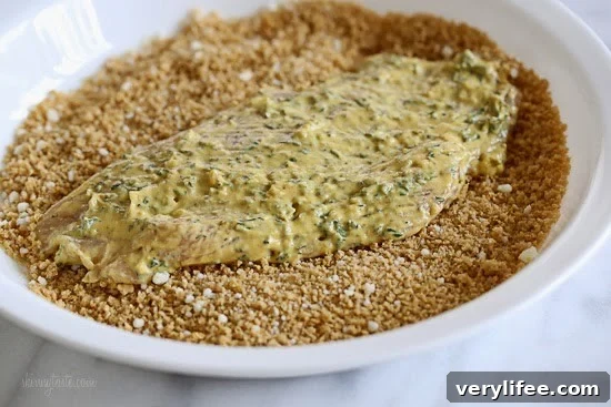 Raw chicken breast covered in dijon mustard with herbs, placed on a plate with breadcrumbs.