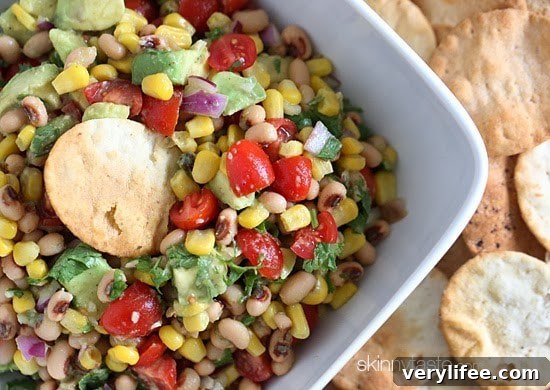 A bowl of black-eyed peas mixed with tomatoes, corn, avocado, and fresh cilantro with a cracker dipping into the bowl