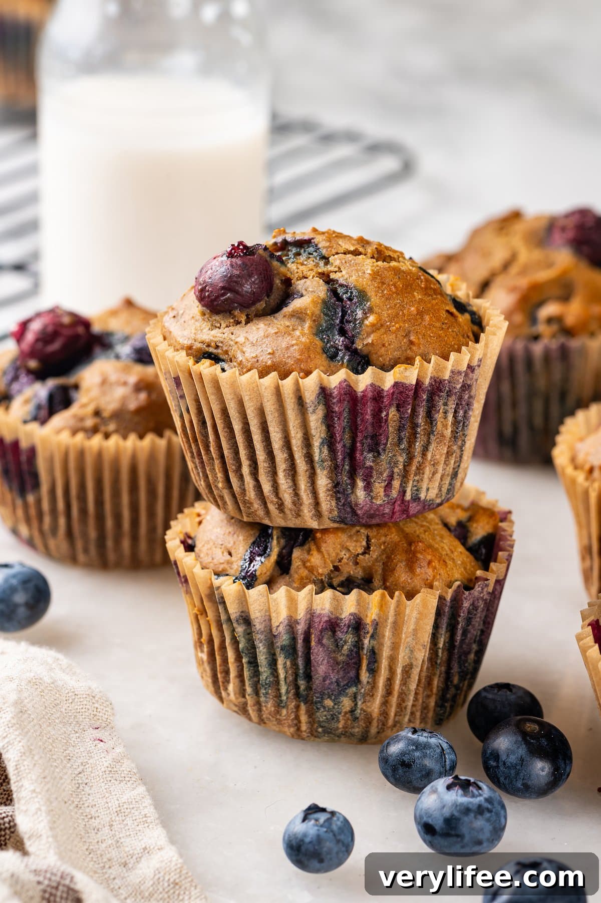 A classic blueberry muffin recipe lightened up with whole wheat flour and applesauce in place of the oil. So moist and delicious! Stack of 2 blueberry muffins with additional muffins and jug of milk in background