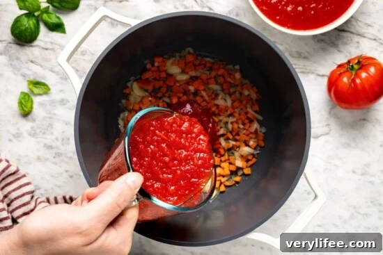 Pouring crushed tomatoes into pot with carrots and onion.