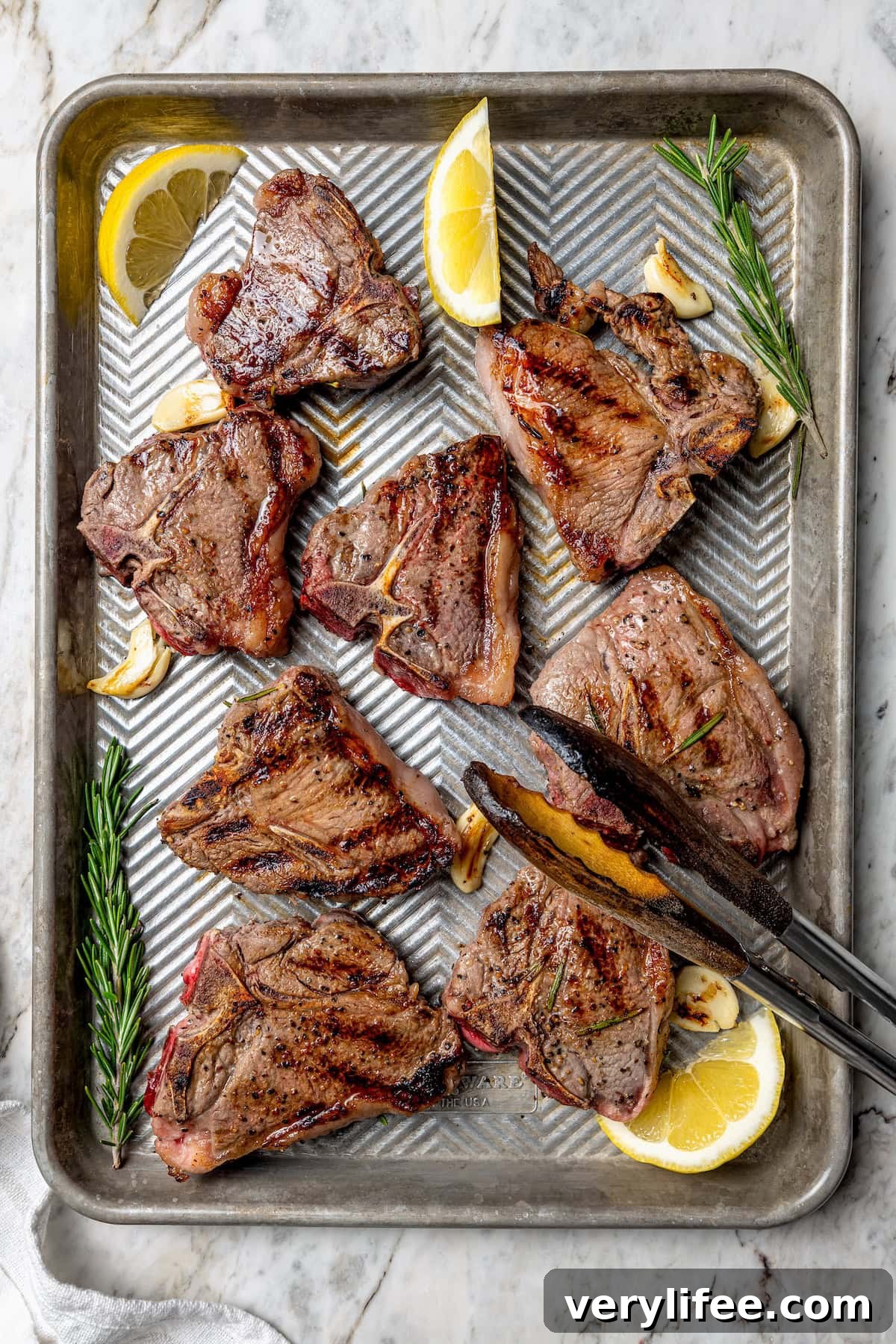 Grilled Rosemary Lamb Chops Recipe with Garlic and Lemon 10 Overhead view of grilled lamb chops on sheet pan with fresh rosemary, lemon wedges, and garlic cloves