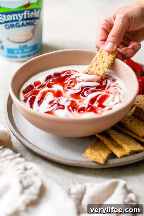 A hand dipping a graham cracker into a bowl of cheesecake dip swirled with strawberry preserves.